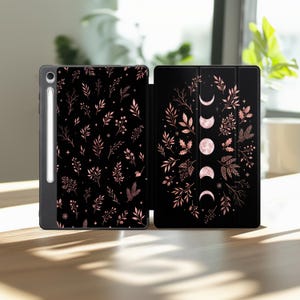 Könnte beinhalten: Eine schwarze Tablet-Hülle mit einem roségoldenen botanischen Muster aus Blättern und Zweigen. Die Vorderseite zeigt eine vertikale Abfolge von Mondphasen, umgeben von einem kreisförmigen Blattwerk, mit einem seitlich befestigten Stift.