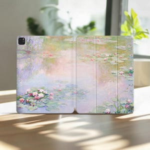 Può includere: Custodia per tablet aperta con un dipinto dai colori pastello di uno stagno di ninfee. L'opera raffigura ninfee rosa, un piccolo ponte e riflessi sull'acqua. La custodia è su una superficie di legno.