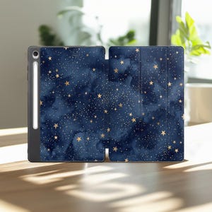 Può includere: Una custodia per tablet blu scuro con un motivo a cielo notturno in stile acquerello, caratterizzata da stelle dorate sparse e minuscoli punti bianchi. La custodia è aperta e rivela un sottile stilo bianco in un alloggiamento verticale.