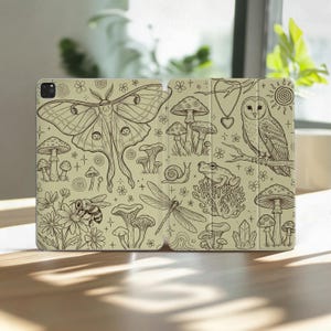 Puede incluir: Una funda para tableta de color beige claro con un patrón detallado de líneas negras que muestra una polilla grande, una abeja sobre una flor, varios hongos, un caracol, una rana, una libélula, un collar con dije de corazón, un sol y cristales.
