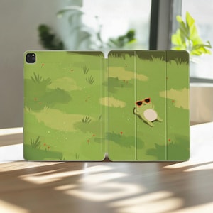 Puede incluir: Una funda para tableta de color verde con una ilustración impresa de una pequeña rana con gafas de sol rojas, tumbada boca arriba en un campo de hierba con flores rojas dispersas. La funda tiene un diseño plegable y un hueco para la cámara.
