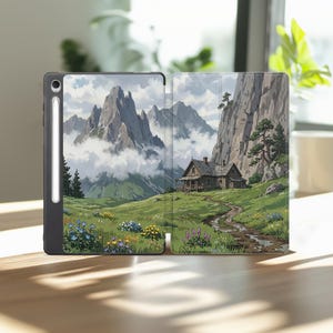 May include: A black tablet case featuring a printed illustration of a wooden cabin nestled in a green mountain valley with a dirt path, wildflowers, and towering rocky peaks partially obscured by clouds, with a stylus docked on the side.