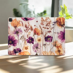 以下が含まれることがあります： 花柄のタブレットケース。紫、オレンジ、黄色の色合いの押し花が白い背景にデザインされています。デザインには、さまざまな種類の花と乾燥した草が含まれています。ケースは開いています。