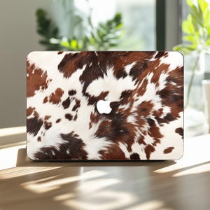 Puede incluir: Funda para portátil con diseño de piel de vaca en tonos marrón, blanco y negro. El logotipo de Apple es visible. La funda está diseñada para adaptarse a un portátil, ofreciendo una capa decorativa y protectora.
