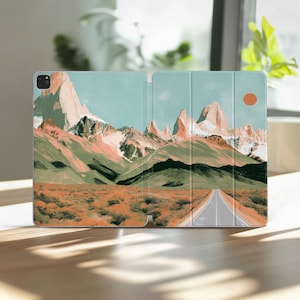May include: An open tablet case featuring a landscape design. The artwork depicts a road leading towards snow-capped mountains under a blue sky with an orange sun. The foreground shows a field of orange and green vegetation.