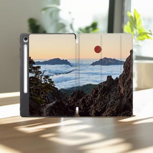 Può includere: Una custodia per tablet nera con una stampa paesaggistica di montagne rocciose che si ergono sopra un mare di nuvole al tramonto con un sole rosso. La custodia include uno slot verticale sul lato sinistro che ospita una penna stilo bianca.