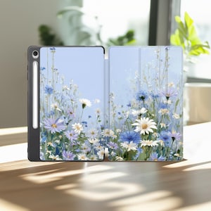 May include: A tablet case with a floral design. The case is open, revealing a light blue background adorned with white, blue, and purple flowers. A black stylus is attached to the side of the tablet. The case is on a wooden surface.