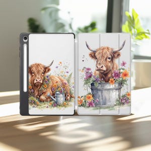 May include: A tablet case featuring a watercolor illustration of a Highland cow. The cow is surrounded by colorful flowers and greenery. The case is open, revealing the tablet screen and a stylus. The design is on a white background.