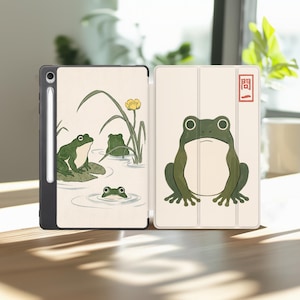 May include: A tablet case featuring a cream-colored cover with illustrations of green frogs in a pond with reeds and a yellow flower. The front panel displays a large centered frog and a red rectangular stamp with the Japanese characters 問一.