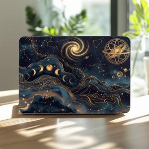 May include: A laptop skin featuring a dark blue celestial design with gold line art. The graphic includes a spiral galaxy, a sun-centered armillary sphere, constellations, a moon phase cycle, and stylized mountain ranges with flowing patterns.