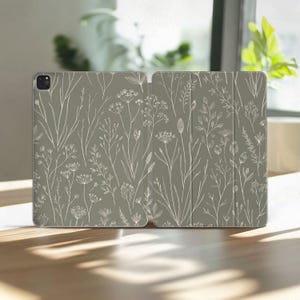 May include: A sage green tablet case featuring a delicate, white botanical illustration. The design includes various wildflowers and foliage, creating a natural and elegant aesthetic. The case is open, showcasing the interior.