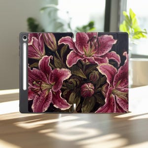 May include: A black tablet case featuring a vibrant floral print of dark pink lilies with green leaves and water droplets on a black background. The case includes a side slot holding a white stylus pen, visible on the left edge of the device.