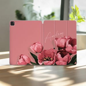 May include: A pink tablet case with a floral design featuring pink tulips and green leaves. The name "Audrey" is written in cursive on the cover. The case is open, revealing the tablet screen and the inside cover.