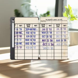 May include: A black tablet case featuring a vintage library due date card design with columns for date due and returned dates. The text reads THE UNIVERSITY LIBRARY DUE DATE and includes various stamped dates from the 1970s and 1980s. A stylus is docked on the left side.