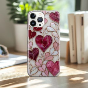 May include: A clear phone case featuring a stained-glass design of hearts in shades of red, pink, and white. The hearts are outlined in gold, creating a romantic and elegant aesthetic. The case is on a wooden surface.