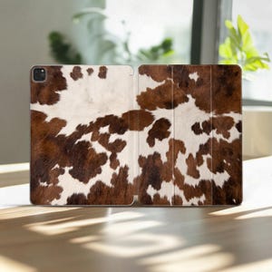 Könnte beinhalten: Eine Tablet-Hülle mit einem braunen und weißen Kuhfellmuster. Die Hülle ist geöffnet und zeigt das Innere. Das Design ahmt das Aussehen von natürlichem Tierfell nach, mit unregelmäßigen braunen Flecken auf weißem Grund. Die Hülle liegt auf einer Holzoberfläche.