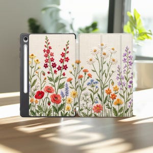 May include: A tablet case with a white textured background featuring an embroidered-style print of colorful wildflowers including red poppies, white daisies, yellow blossoms, and purple stalks, with a stylus pen docked in the side slot.