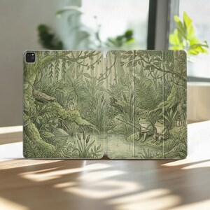 Puede incluir: Funda de tableta abierta con una ilustración de un bosque. Dos ranas de dibujos animados pescan junto a un estanque. La obra de arte es principalmente verde y beige, con representaciones detalladas de árboles, plantas y una linterna.