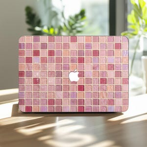 May include: A laptop with a pink mosaic tile design. The cover features a pattern of small, square tiles in shades of pink, rose, and mauve. A white Apple logo is centered on the cover. The laptop is on a wooden surface.