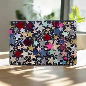 Pode incluir: Uma capa de tablet azul-escura com um padrão denso de estrelas variadas em diversas texturas, cores e acabamentos, incluindo veludo vermelho, glitter dourado, efeitos holográficos, ganga e padrões de espiral a preto e branco em toda a superfície.