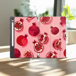 May include: A tablet case with a light pink background featuring a repeating pattern of whole and halved red pomegranates. The case has a black frame and a dedicated vertical slot on the left side containing a white stylus pen.