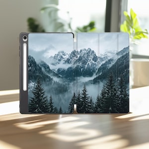May include: A black tablet case featuring a printed landscape of snow-capped mountains, dense evergreen forests, and low-hanging mist. The case includes a dedicated vertical slot on the left side for a stylus, which is currently docked in place.