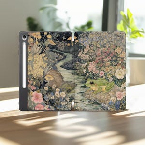 May include: A tablet case featuring a watercolor illustration of a serene landscape with a stream, flowers, and a green frog. The case is open, revealing the screen and a stylus. The color palette includes greens, pinks, and yellows.