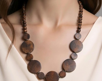May include: A necklace featuring dark brown wooden beads of varying sizes arranged in a circular pattern. The design includes smaller spherical beads along the upper sections and larger, flat circular wooden discs suspended towards the centre.