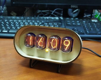 Pode incluir: Um relógio de madeira com quatro tubos Nixie que mostram a hora 16:09. O relógio tem um acabamento marrom claro e uma forma retangular arredondada. Os tubos Nixie brilham em laranja.