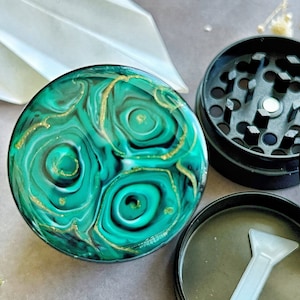 Pode incluir: Um moedor de resina verde e dourada com design de malaquite. O moedor é redondo e tem uma parte superior e inferior pretas. O moedor está desmontado, mostrando os componentes de moagem e um pequeno raspador.
