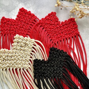 May include: Close-up of handmade macrame pieces in red, cream, and black. The red pieces are the largest, with a textured, knotted design and long fringe. The cream and black pieces have a similar design. Decorative craft items.