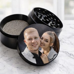 May include: Black, round herb grinder with a lid featuring a color photo of a smiling couple. The grinder is open, revealing a mesh screen and the grinding teeth. The product is designed for herbs.