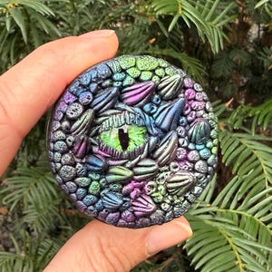 Pode incluir: Uma escultura redonda e iridescente de um olho de dragão com íris verde e pupila preta. A escultura apresenta escamas texturizadas em tons de azul, roxo e verde. A obra de arte é segurada em uma mão contra um fundo de folhagem verde.