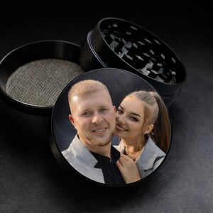 Pode incluir: Moedor de ervas de metal preto com uma foto personalizada na tampa. A foto mostra um casal sorrindo. O moedor está aberto, revelando a tela de malha e os dentes de moagem. O fundo é escuro.