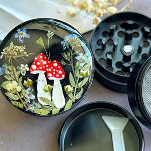 Pode incluir: Um moedor preto com uma tampa transparente que apresenta um design de arte em resina de cogumelos vermelhos e brancos e flores prensadas sobre um fundo preto.