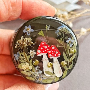 May include: A round, black resin object with a whimsical design. Two red and white mushroom figures are surrounded by dried flowers and greenery. The resin is clear, allowing a view of the embedded botanical elements.