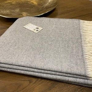 May include: A grey throw blanket with a white fringe. The blanket is folded in half and has a small white tag attached to it.