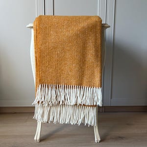 Può includere: Una coperta a motivo a spina di pesce giallo senape con frange bianche è drappeggiata su una sedia bianca. La trama e il colore della coperta creano un'estetica calda e invitante. Le frange aggiungono un elemento decorativo.