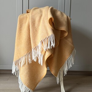 May include: A mustard yellow throw blanket featuring a subtle herringbone weave pattern and long, white twisted fringe trim along the edges. The fabric has a soft, textured appearance and is draped to show its folds and layered construction.