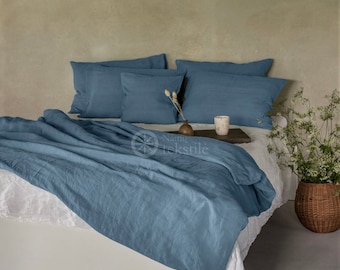 May include: A blue linen duvet cover with a white linen sheet underneath. The duvet cover is folded back to reveal the sheet. There are three blue linen pillows on the bed, with a small vase and a cup on a wooden tray on top of the pillows.