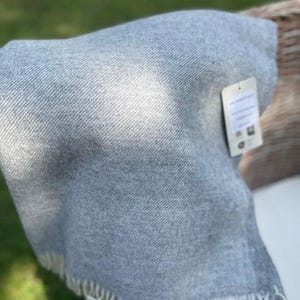 May include: A folded, pale grey blanket with a fringed edge. The blanket has a woven texture and a small tag attached. The fringe is cream-coloured. The blanket is draped over an unseen object.