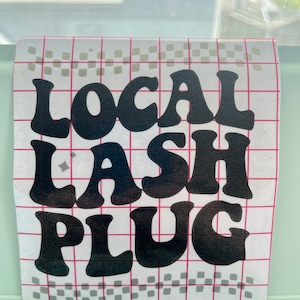 May include: A clear sticker with the words "LOCAL LASH PLUG" in large, black, bubble letters. The sticker has a grid pattern with pink lines and gray squares. The sticker is on a light green surface.