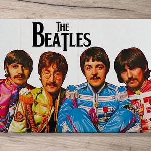 May include: A white phone case with a colorful image of The Beatles. The Beatles are wearing colorful outfits and have their signature hairstyles. The text "The Beatles" is printed in black letters on the case.