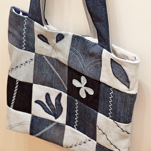 Puede incluir: Un bolso tote de mezclilla patchwork con un diseño floral. La bolsa presenta una mezcla de cuadrados de mezclilla azul claro y oscuro, con detalles bordados de flores y hojas. Las asas son del mismo material de mezclilla.