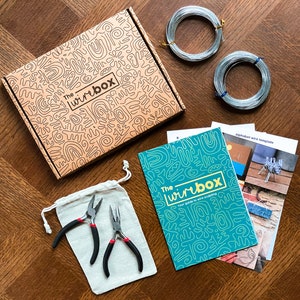 May include: A wire sculpting kit featuring "The Wirbox" branding on the box and booklet. The kit contains two silver wire spools, a drawstring bag with two pliers, and instructional materials. The box has a brown background with a white pattern.