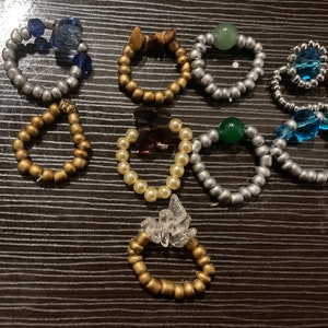 May include: A collection of ten beaded rings featuring a variety of colours and materials, including gold, silver, blue, green, and white beads. Some rings incorporate decorative elements such as crystals and charms.