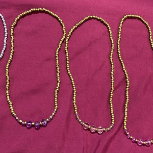 May include: Five beaded necklaces with different coloured beads. The necklaces are made with small, round beads in silver, gold, and a variety of colours. The necklaces are laid out on a pink fabric background.