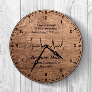 Puede incluir: Un reloj de madera con un gráfico de latido del corazón y el texto "Talented enough to be a cardiologist. Crazy enough to love it." El reloj también tiene el texto "Dr. Jack Walsh, Columbia University, June 2020".