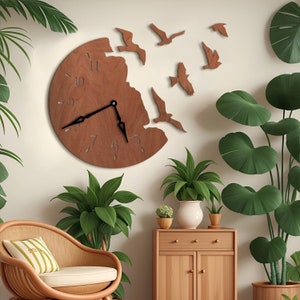Puede incluir: Un reloj de madera con una esfera circular y agujas negras. El reloj está diseñado para parecer que los pájaros están volando fuera de él. El reloj está colgado en una pared blanca con plantas verdes en el fondo.