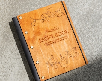 May include: A wooden recipe book with a black leather spine. The cover is engraved with the text "Amanda's Recipe Book", "Love is finding someone worth sharing your cookies with", and "Est. 2023".